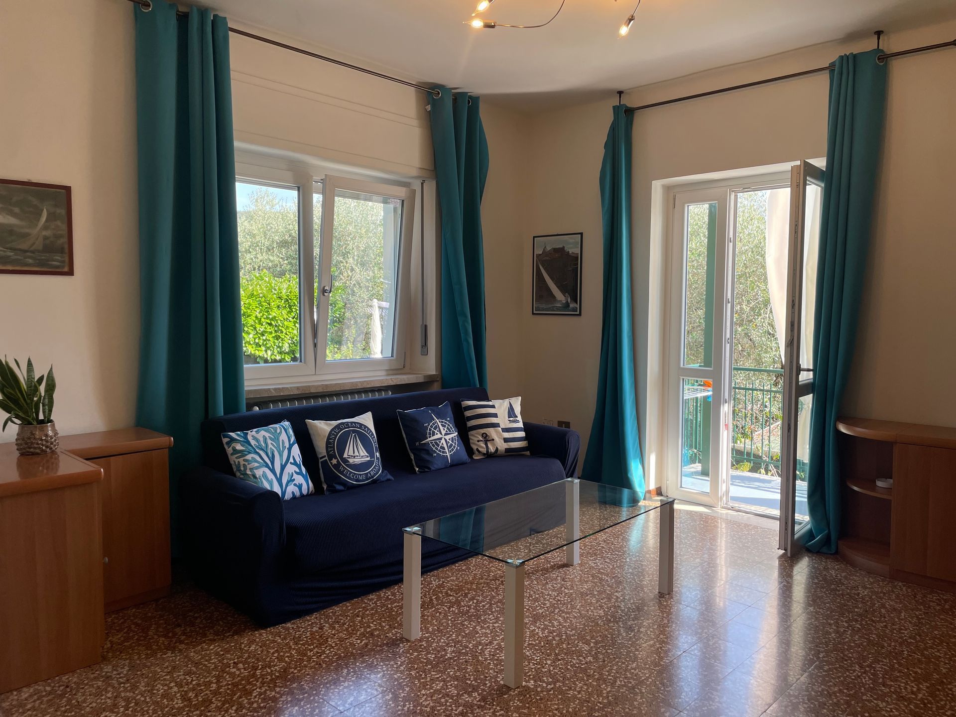 Salotto con divano blu con cuscini marinari appoggiato sotto una finestra con tende blu, sulla sinistra del divano c'è la porta finestra sempre con tende blu che si affaccia sul balcone coperto
