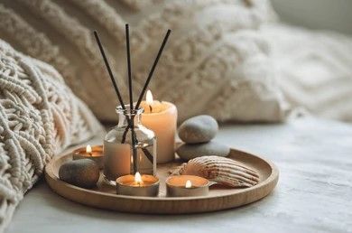 A wooden tray holds a reed diffuser, three lit candles, two smooth grey stones, and a seashell on a soft, textured surface.