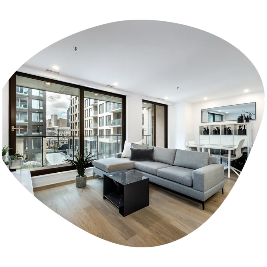 Modern living room with gray sectional, black coffee table, wood floors, and large windows overlooking apartments