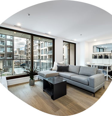 Modern living room with gray sectional sofa, black coffee table, and floor-to-ceiling windows overlooking apartments