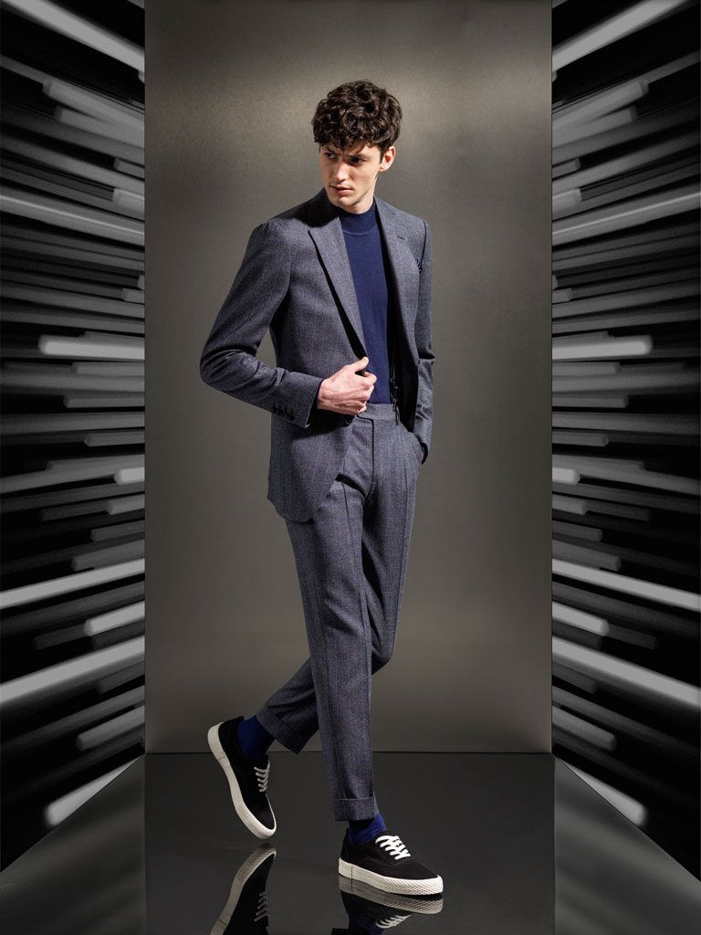 Man in a gray suit and navy turtleneck posing in a studio with a modern black-and-white backdrop