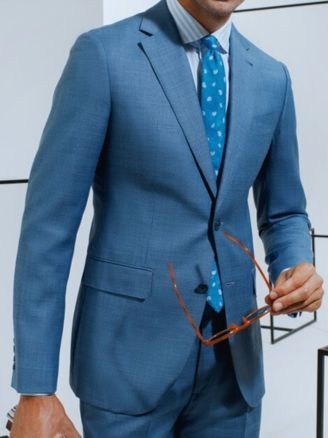 Man in a light blue suit holding glasses, standing against a white wall