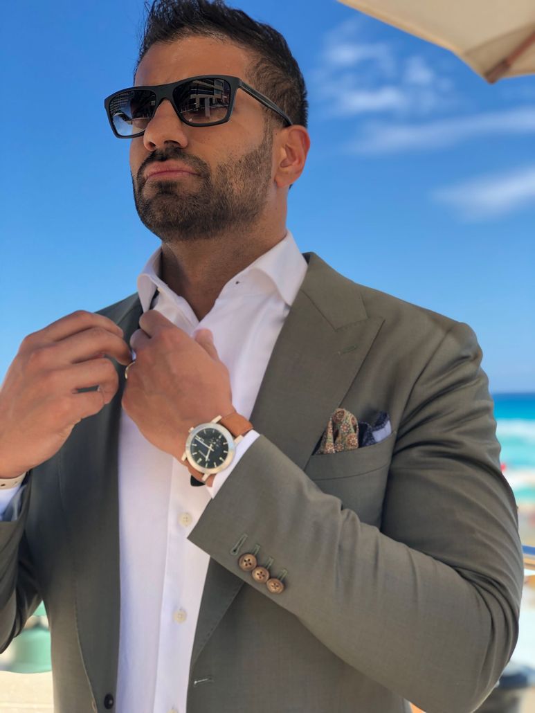 Man in sunglasses adjusting his blazer against a blue sky, wearing a white shirt and wristwatch.