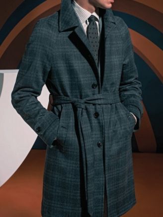 Man wearing a teal plaid overcoat and tie, hands in pockets, standing against an abstract backdrop