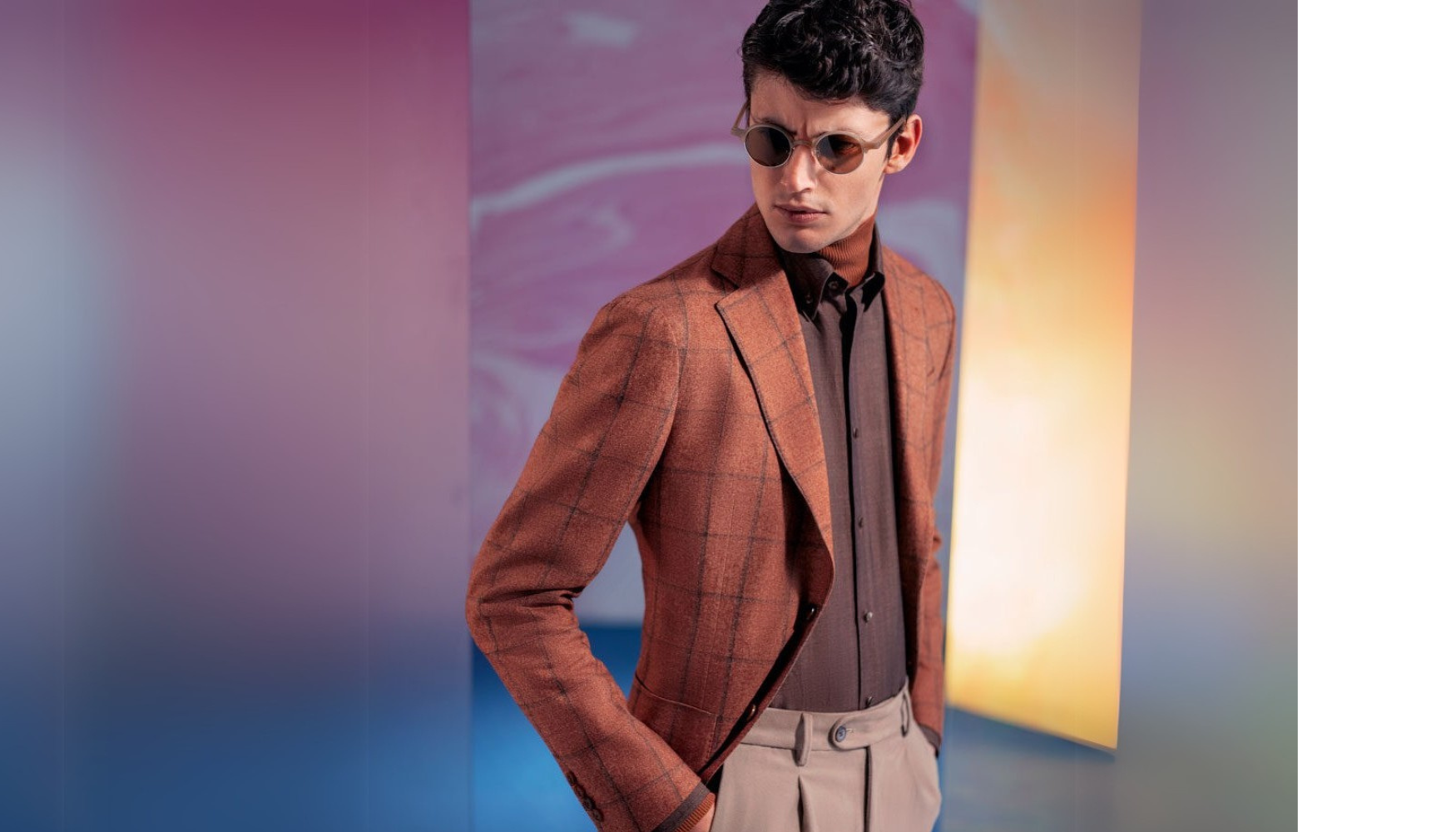 Man posing in a brown blazer, dark shirt, and sunglasses against a purple and orange studio backdrop