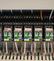 Electrical control panel with rows of black circuit breakers and orange wiring, some labeled green switches.