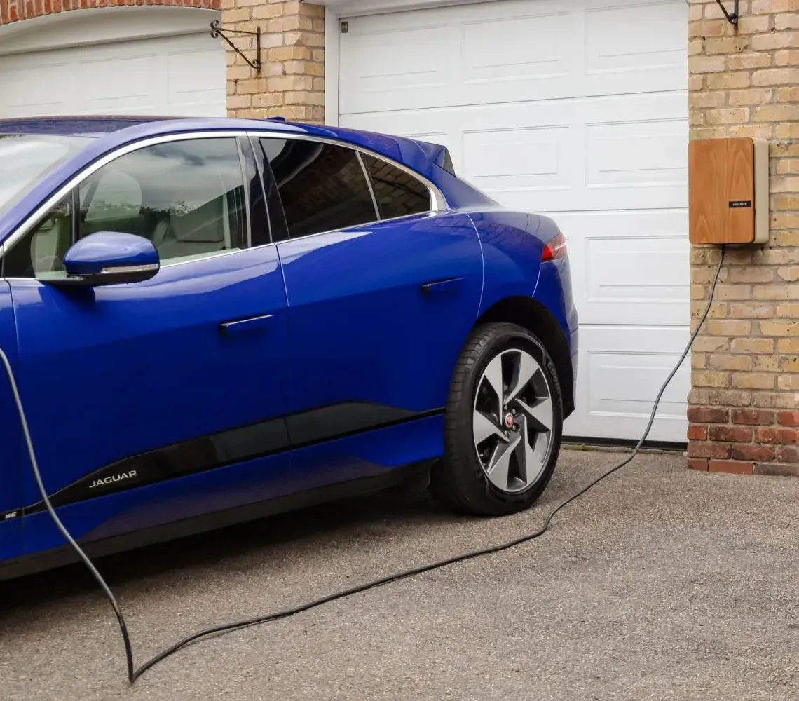 Blue electric car charging outside a garage, with a charging cable plugged in.