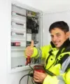 Electrician in a high-visibility jacket gives a thumbs up beside an open circuit breaker panel.