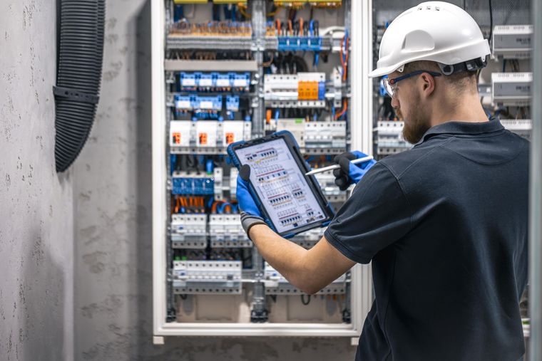 Technician using a tablet to inspect an electrical control panel with wiring and circuit breakers