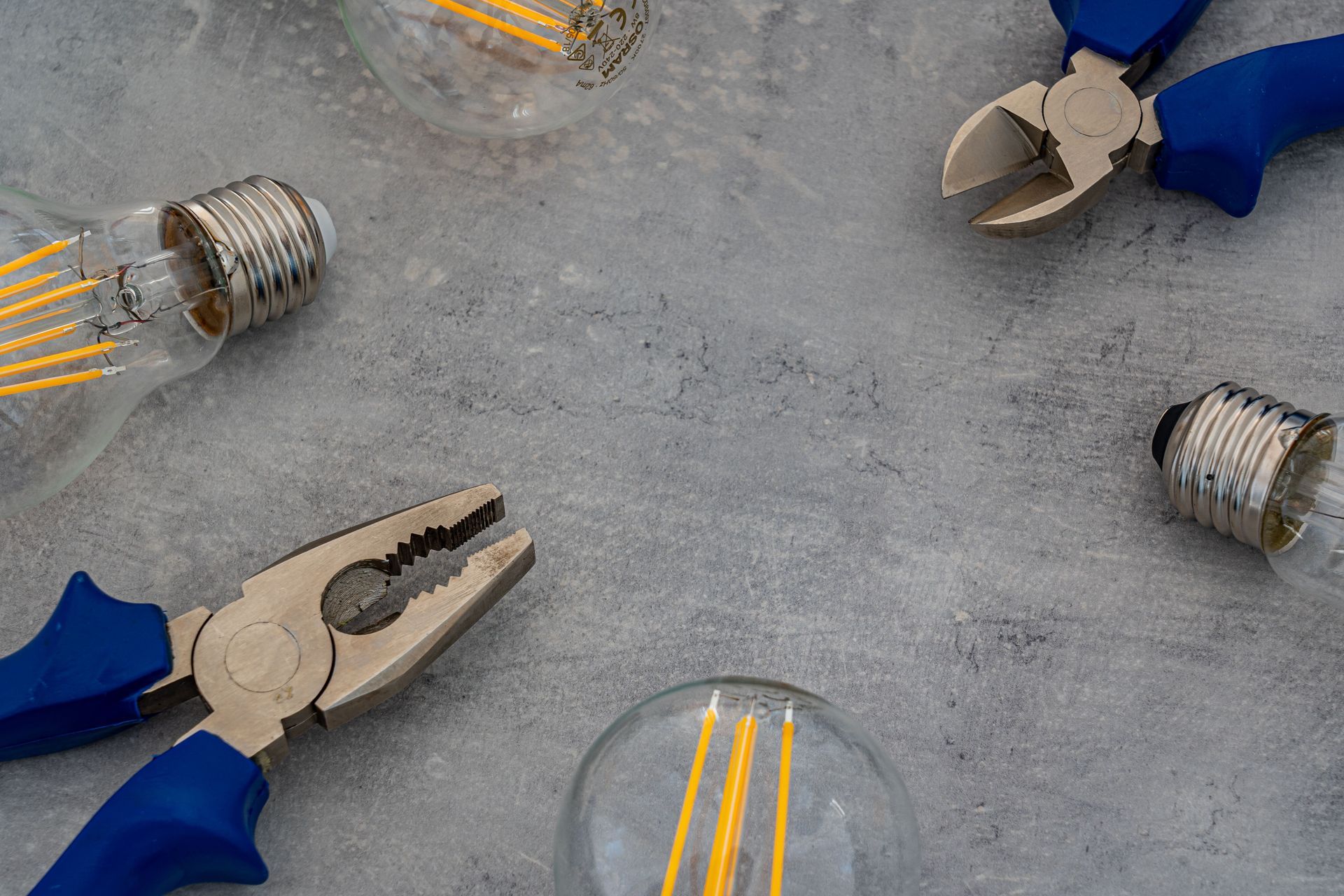 Blue-gloved hands with pliers around scattered light bulbs on a gray surface