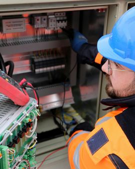 Technician in blue hard hat inspecting an electrical control panel with exposed circuitry