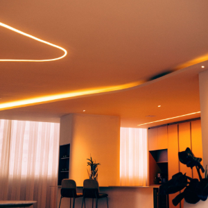 Warm modern room with curved orange ceiling lights, sheer curtains, chairs, and a plant near a window.