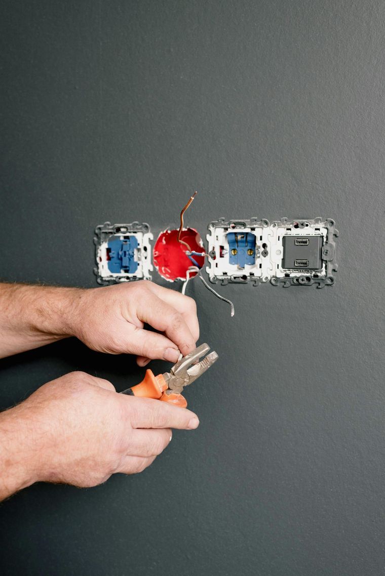 Hands wiring a red electrical box in a wall outlet opening with tools nearby