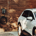 White SUV parked by a wood wall with a wall-mounted EV charger and coiled cable