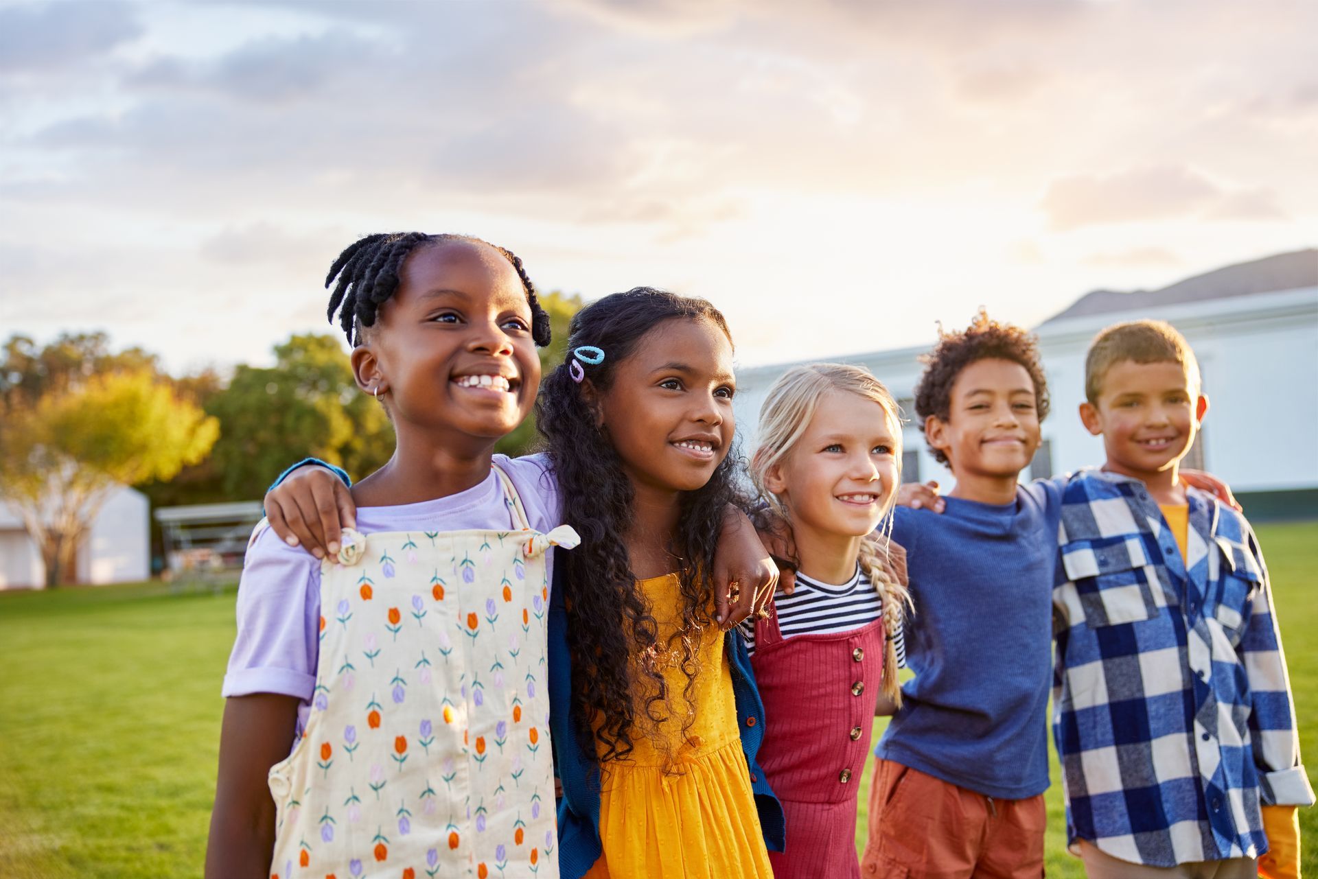 Five diverse children with arms around each other, smiling in a sunny outdoor setting.
