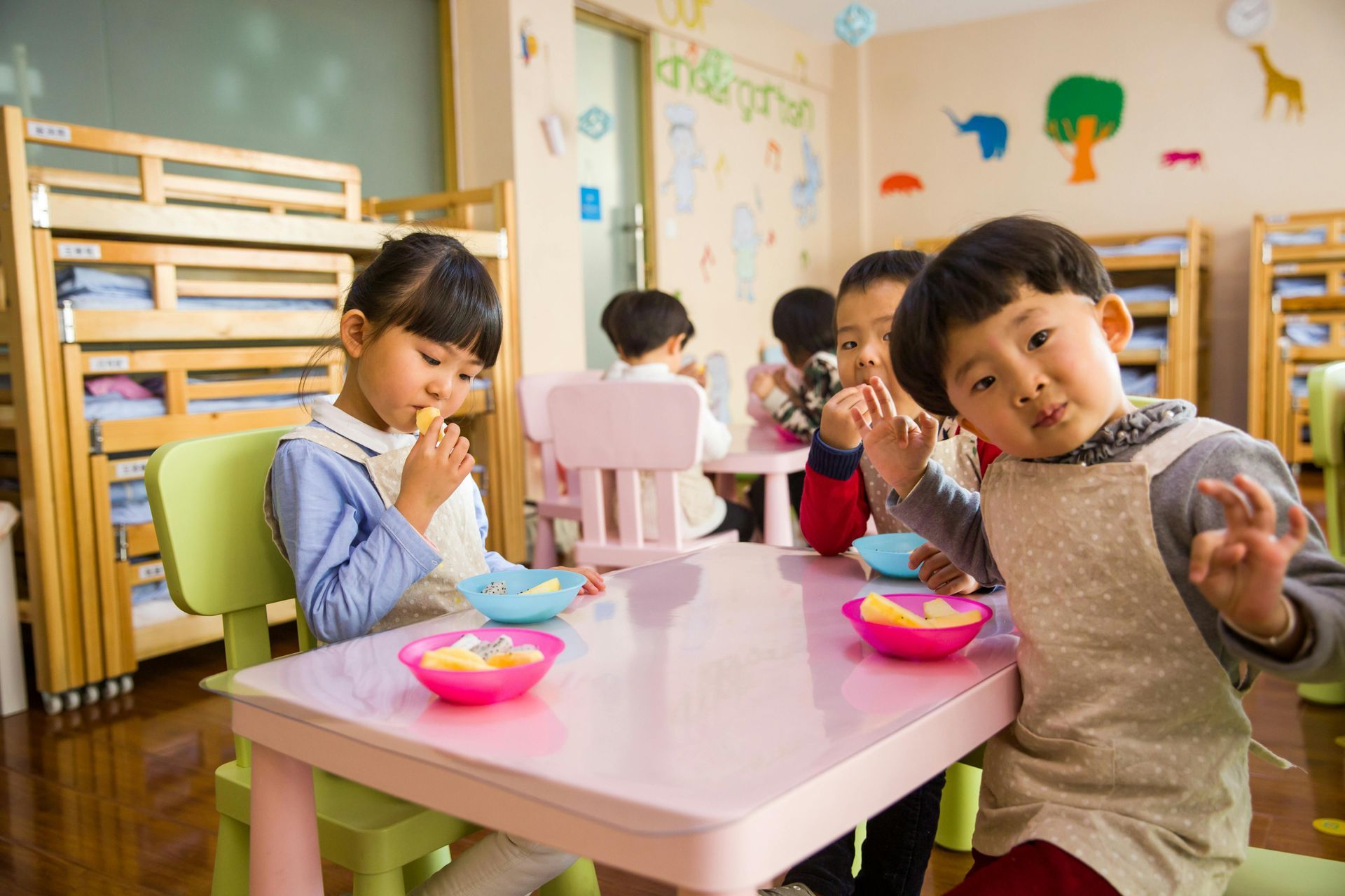 Children at a pink table in a classroom, eating fruit from colorful bowls. One child makes an 