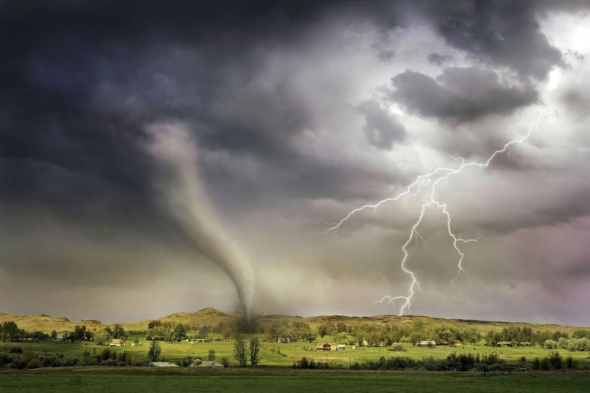 Tornado tearing across a green landscape under dark, stormy clouds with lightning.