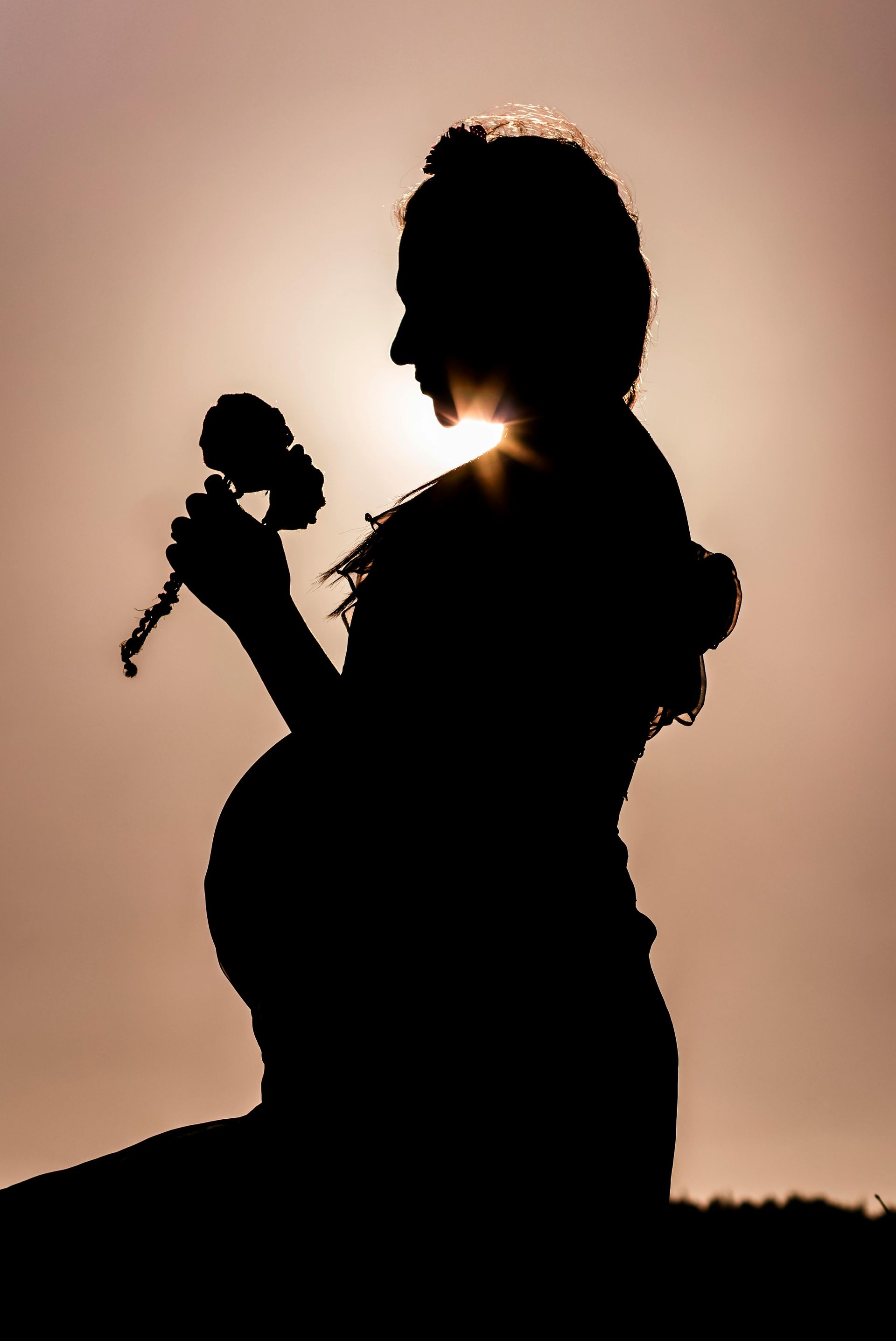 Silhouette of pregnant woman kneeling, holding a flower, with the sun behind her.