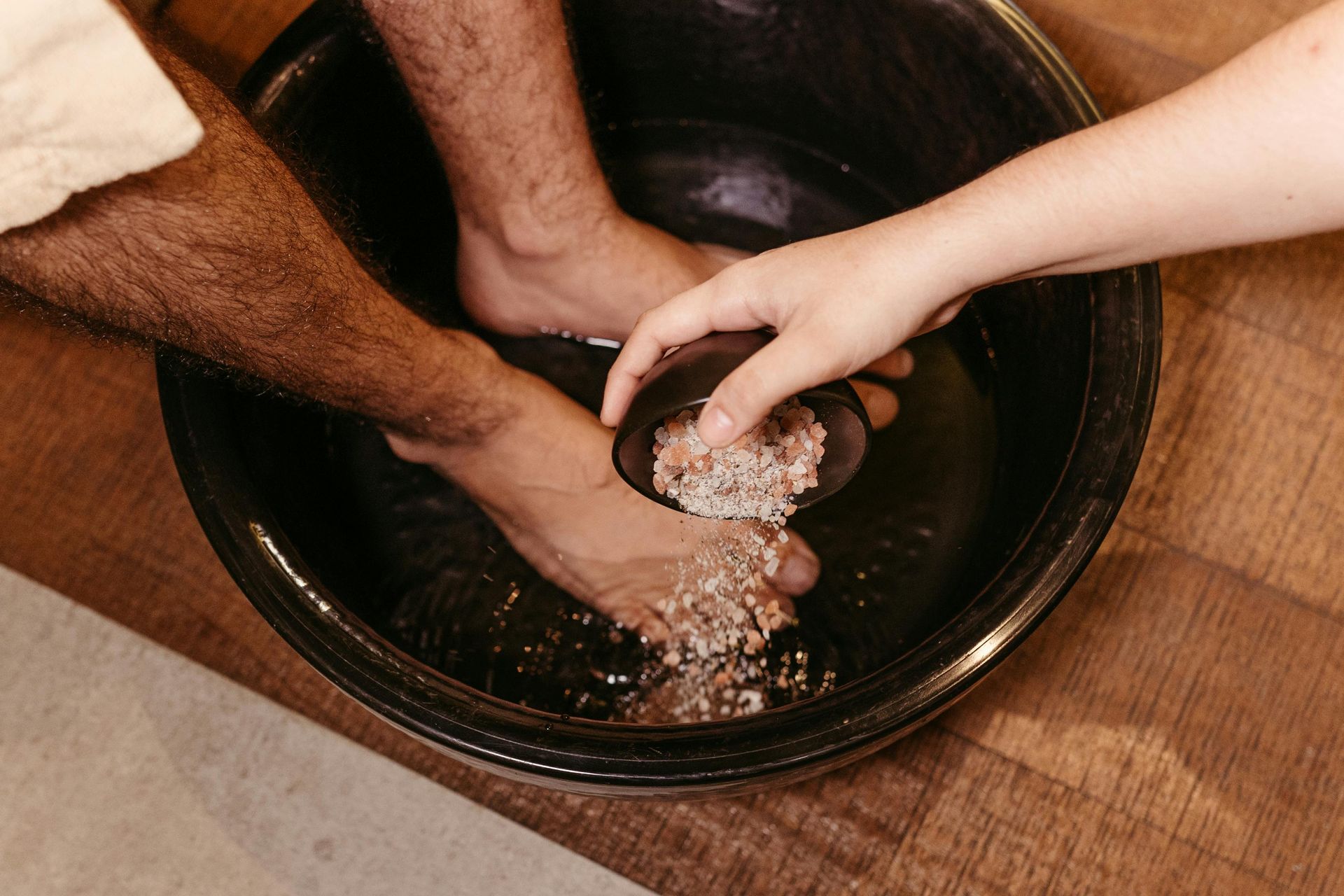 Person's feet in a black bowl being washed by another person. Wooden floor in the background.