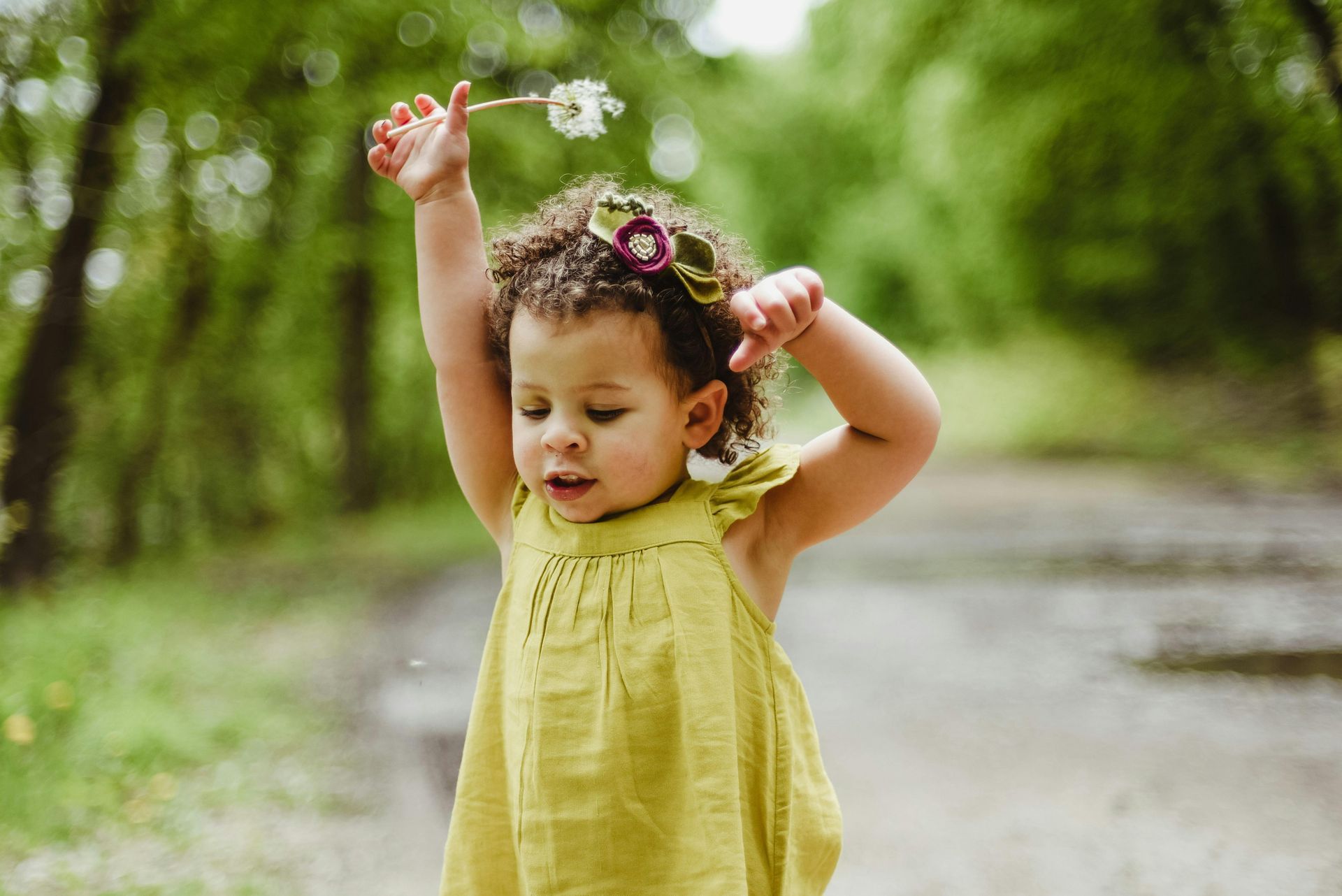 Young child in green dress holds flower, arms raised, on a wooded path.