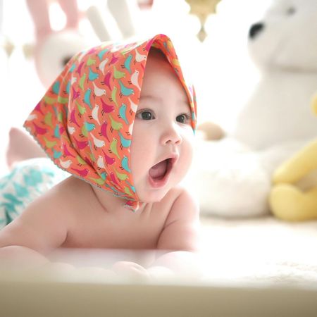 Baby with mouth open wearing a patterned head covering, lying on stomach, surrounded by toys.
