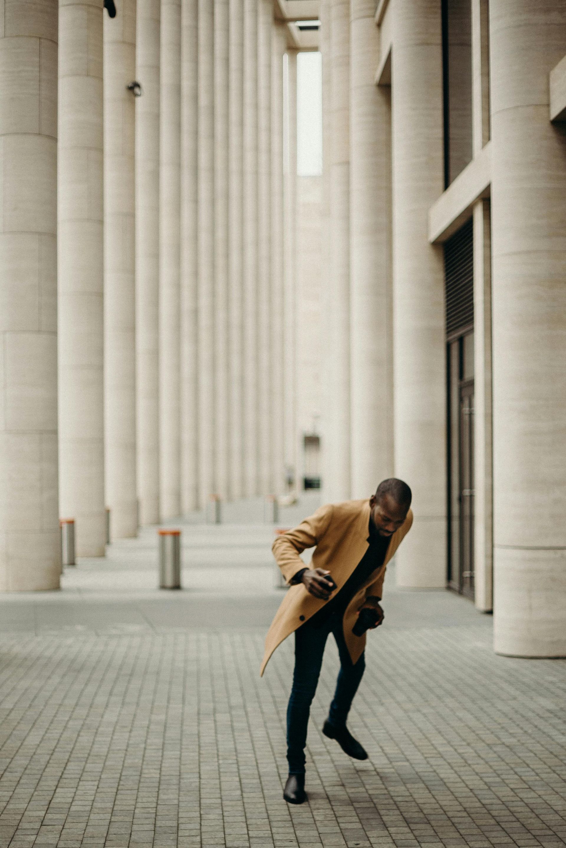 Man in tan coat dancing on a brick path with tall, white columns in the background.