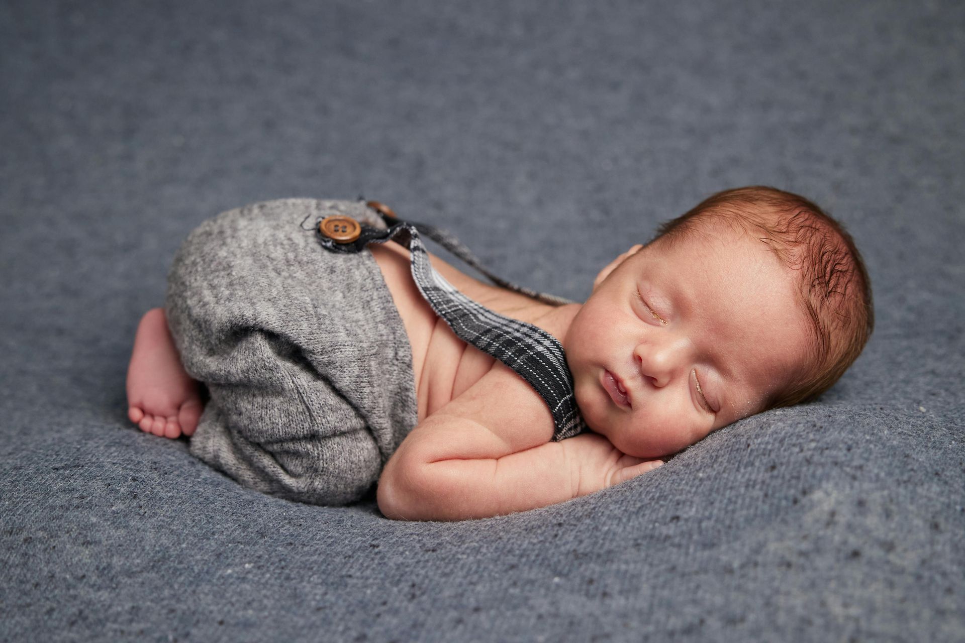 Newborn baby sleeping on a blue blanket, wearing gray suspenders.