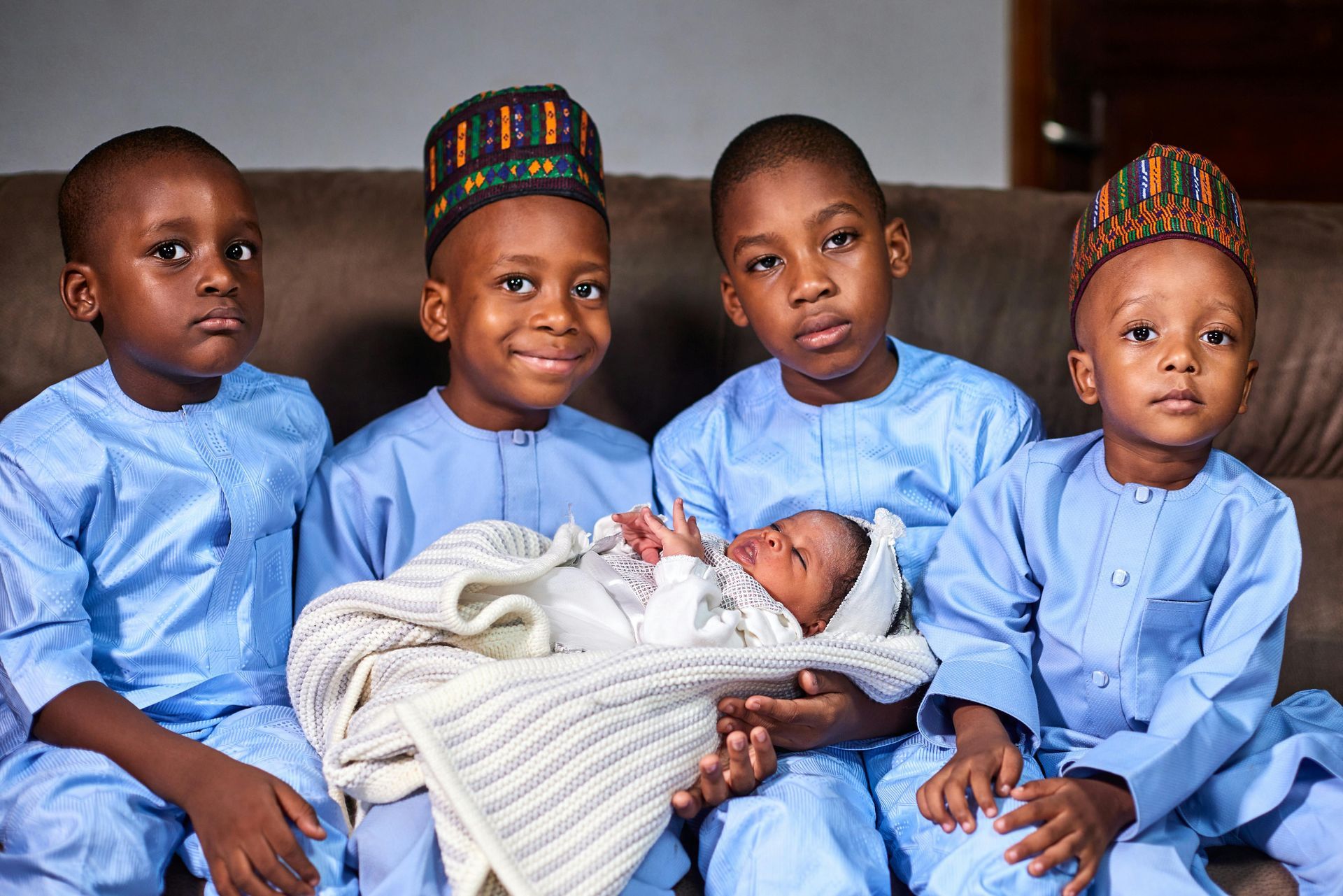 Four boys in blue outfits with a newborn, all seated on a brown couch.