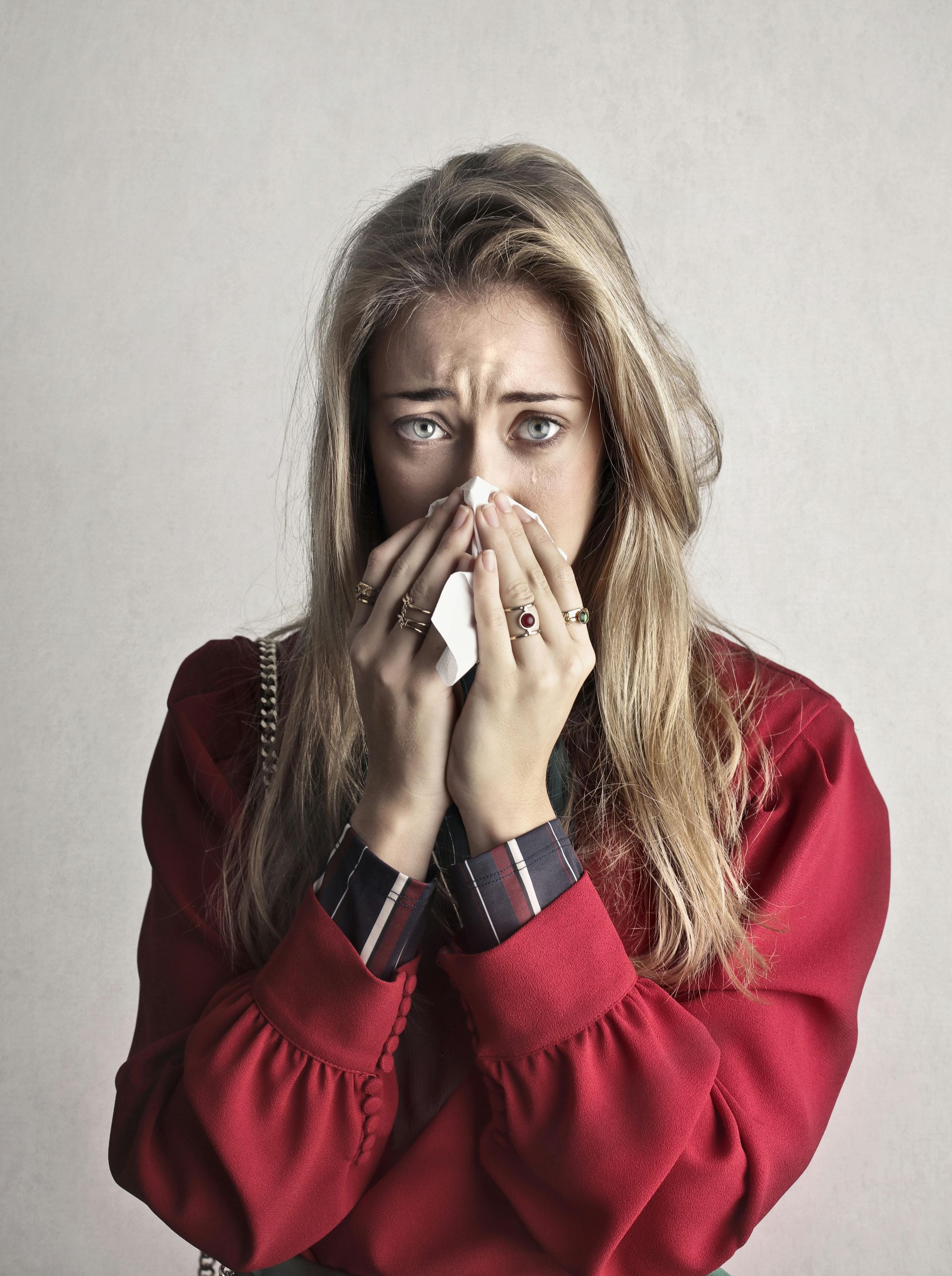 Woman with long blonde hair, in red sweater, blowing her nose with a tissue, looking sad.