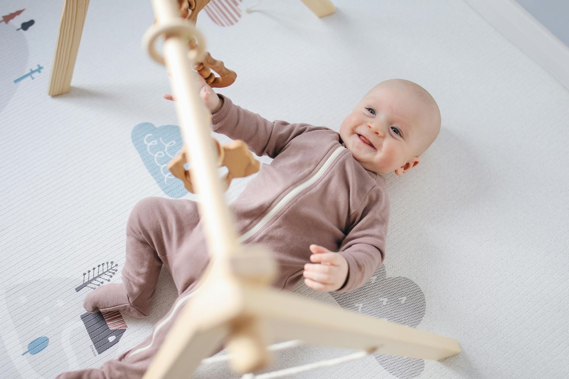 Baby in mauve onesie smiles, reaching for wooden play gym with wooden toys on mat.
