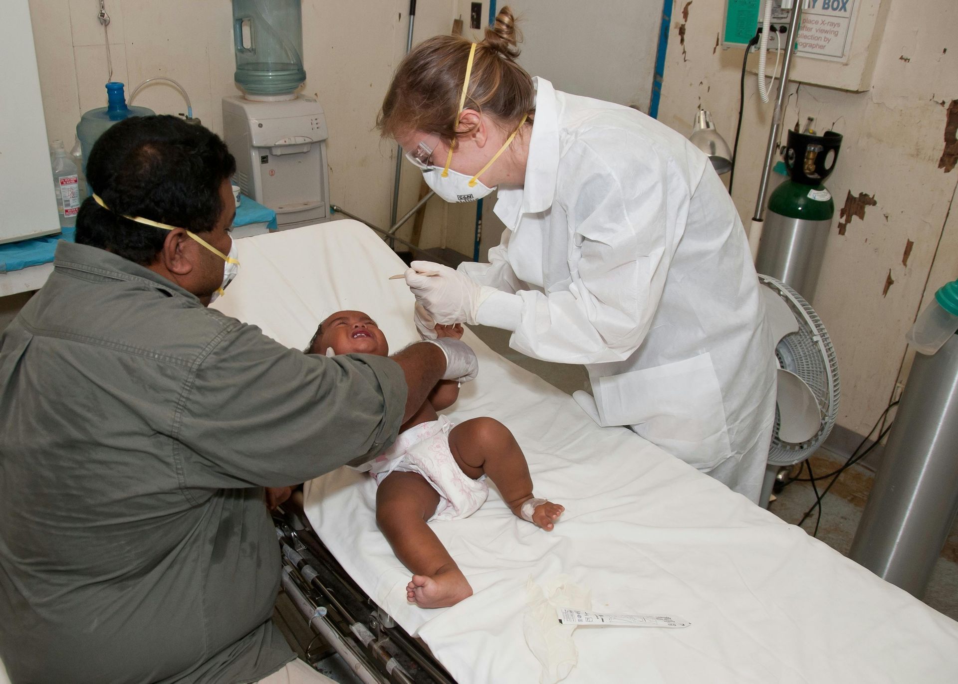 Two adults treat a baby on a bed in a medical setting; both adults wear masks, one in a lab coat.