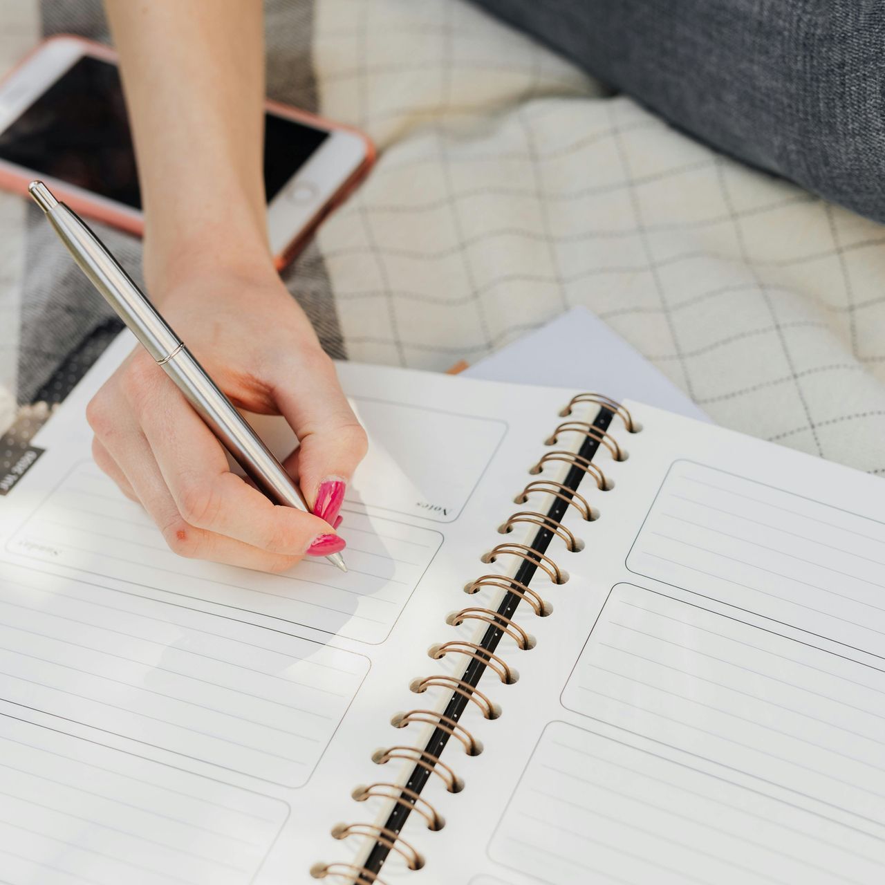 Woman writing with a silver pen in a spiral notebook. Pink nails, setting is outdoors, phone nearby.
