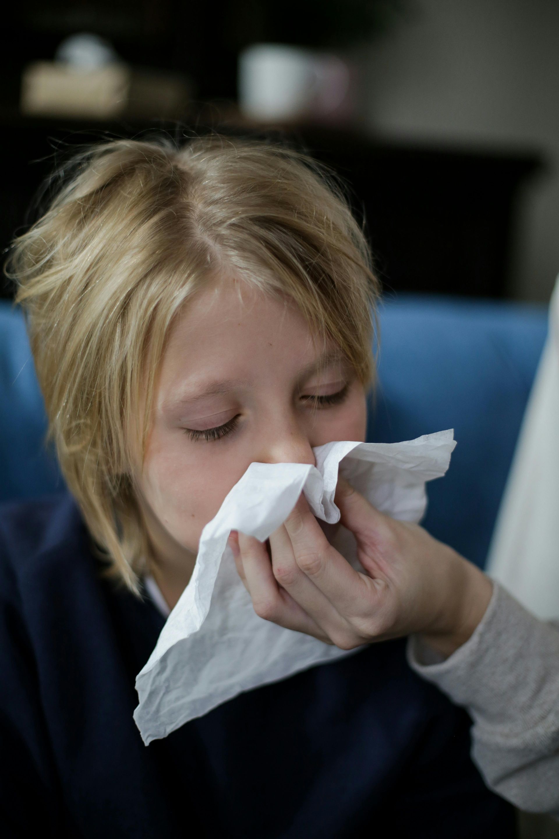 A child with blond hair blowing their nose with a tissue, indoors.