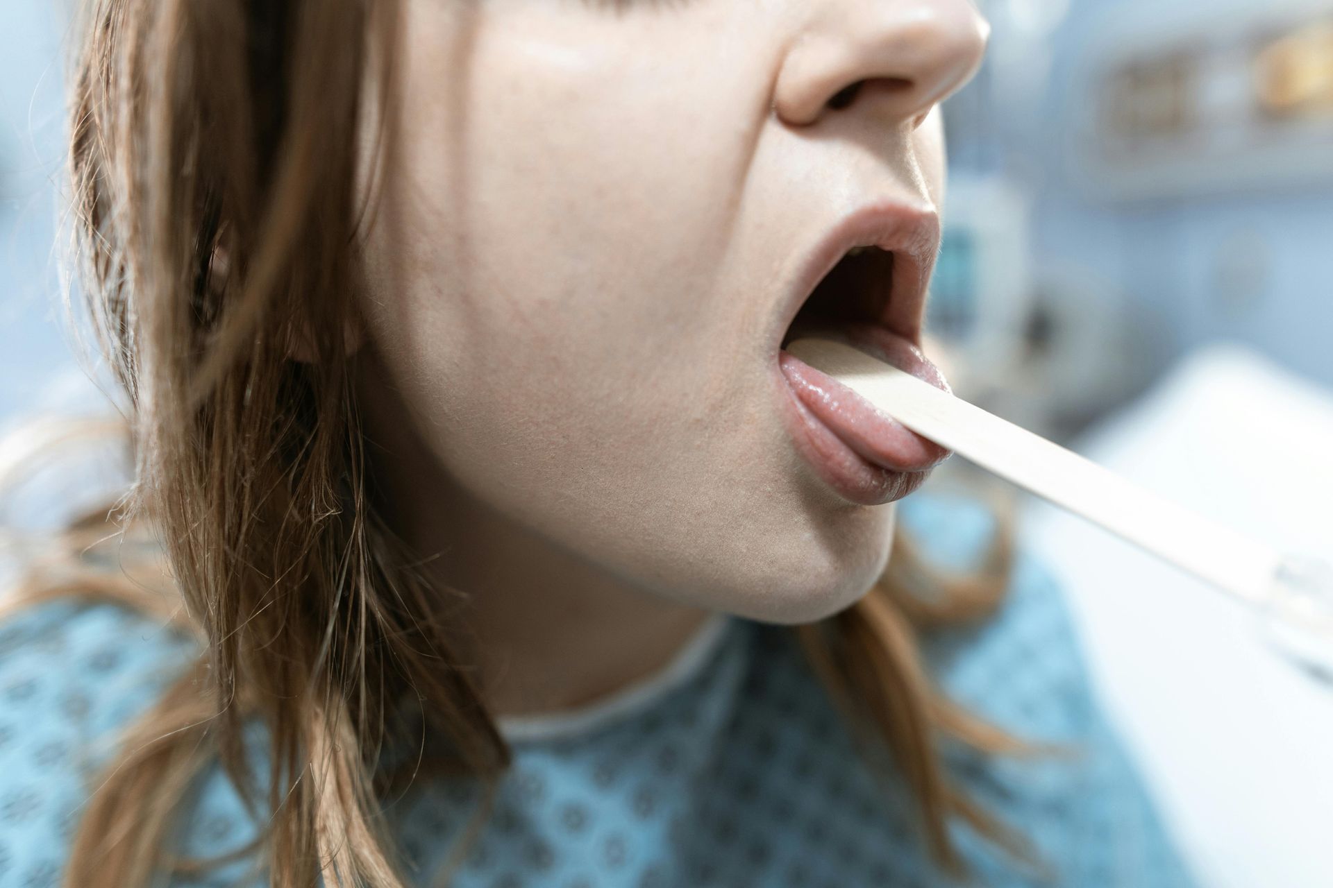 A woman in a blue hospital gown has her mouth open, tongue depressed, during a medical exam.