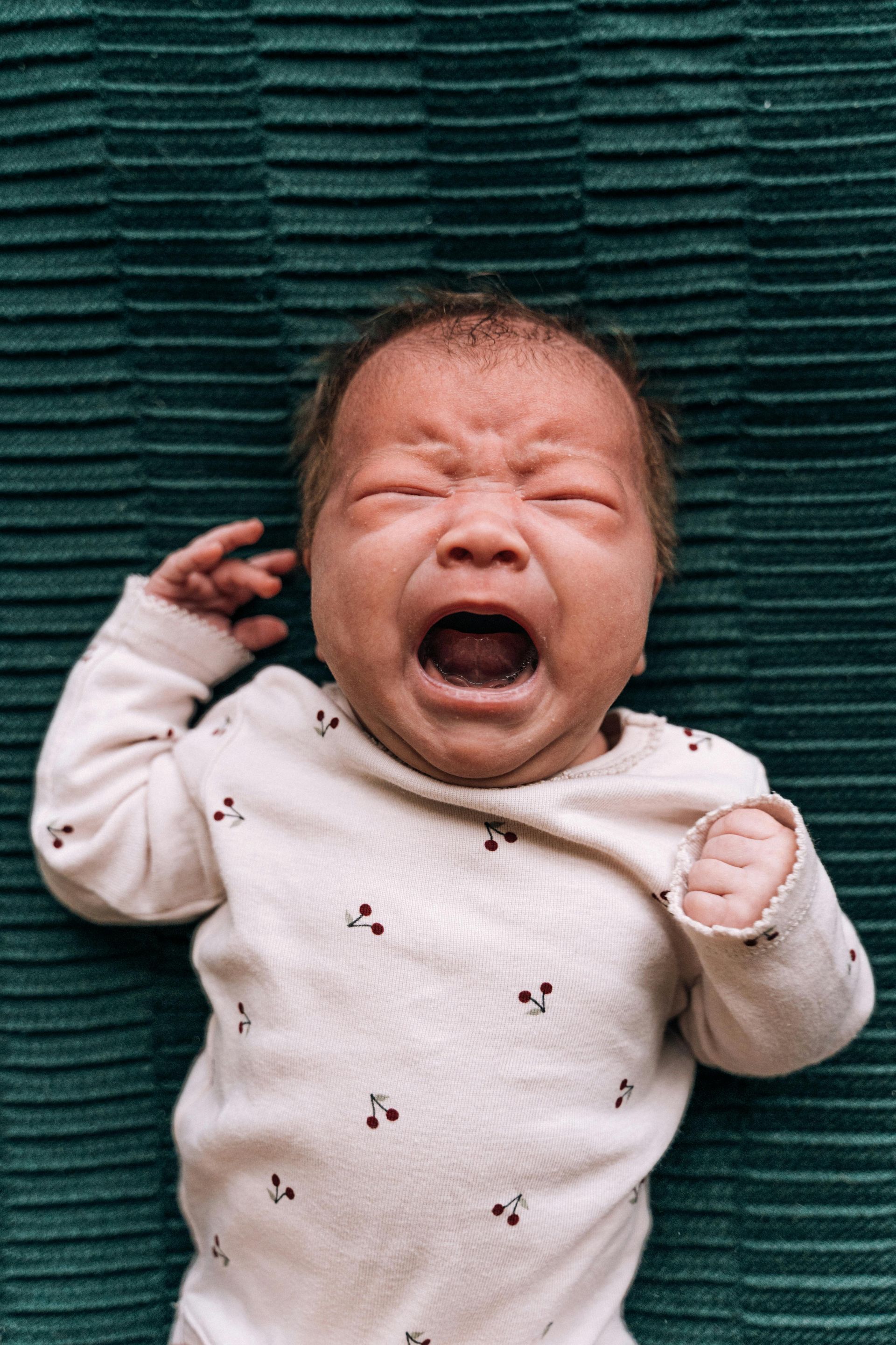 Baby crying, lying on a green textured surface. Wearing a white onesie with red cherry pattern.