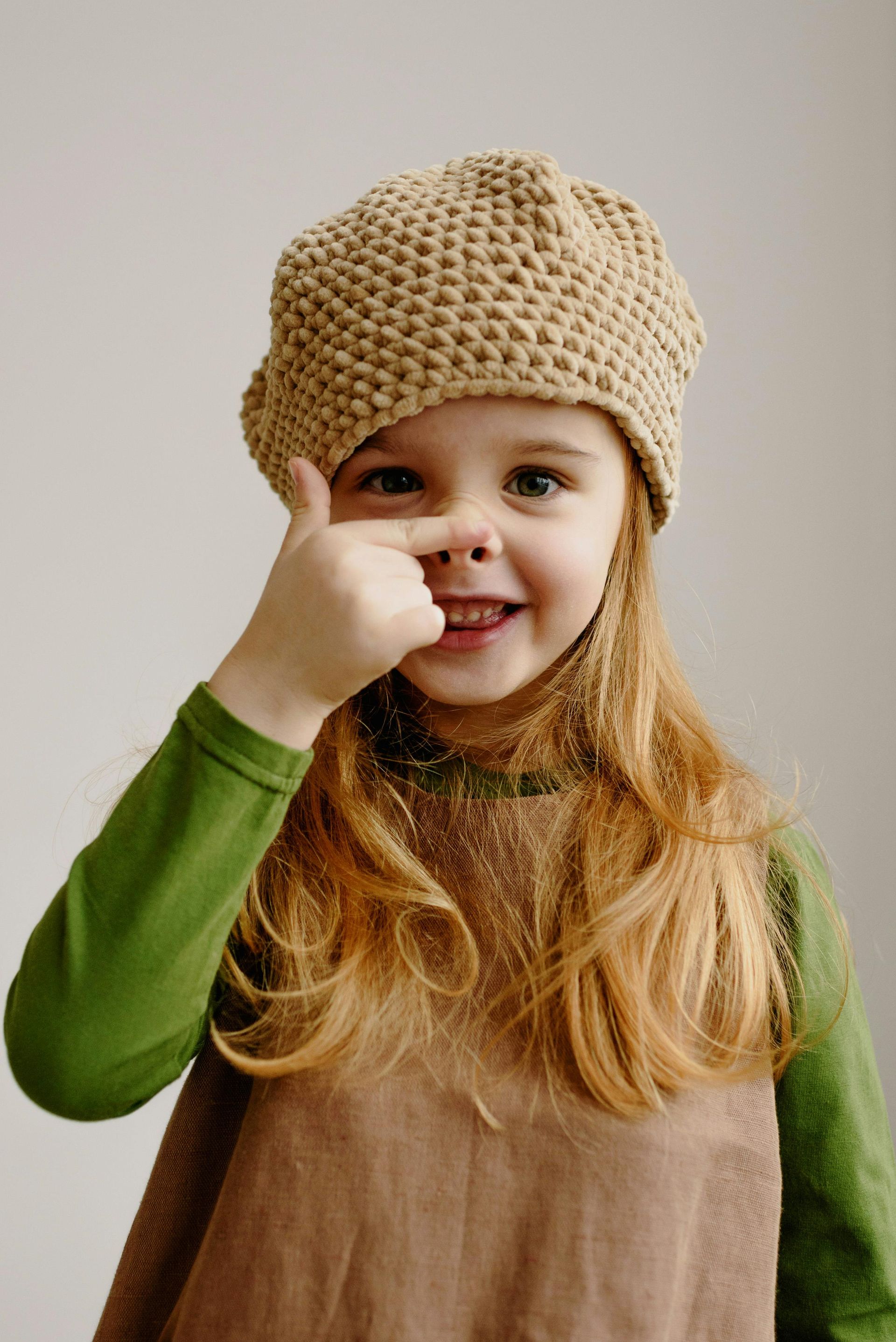 Girl with red hair, wearing a brown beret, smiles while touching her nose.