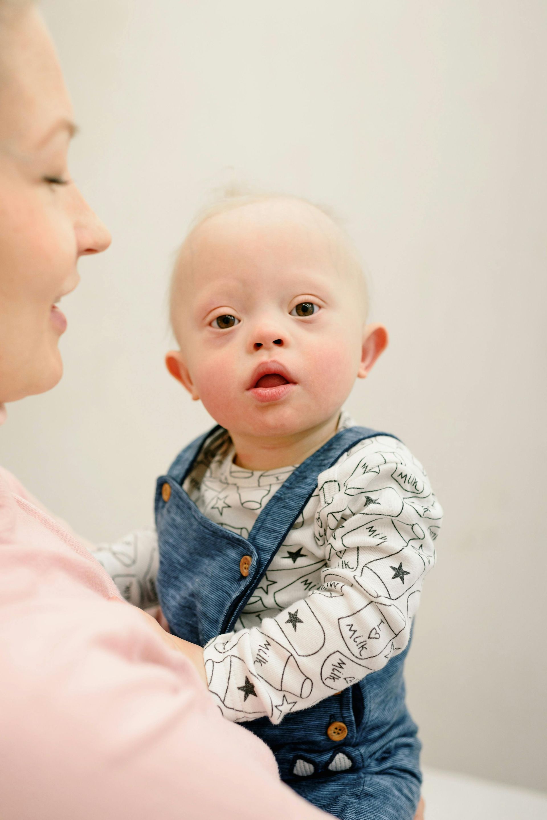 Woman holding a baby with Down syndrome; baby is looking at the viewer, wearing overalls.