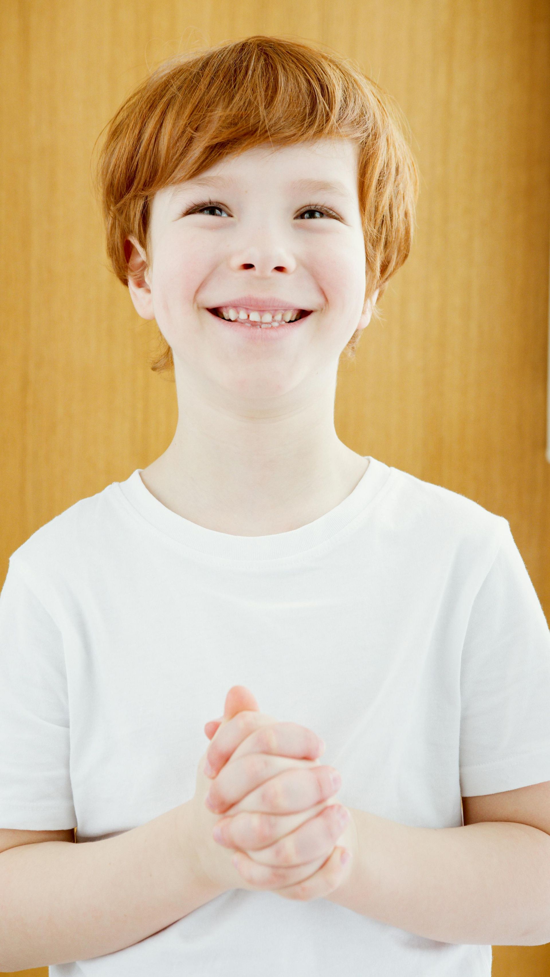 Red-haired boy in white shirt smiles, hands clasped, against a wood-paneled background.