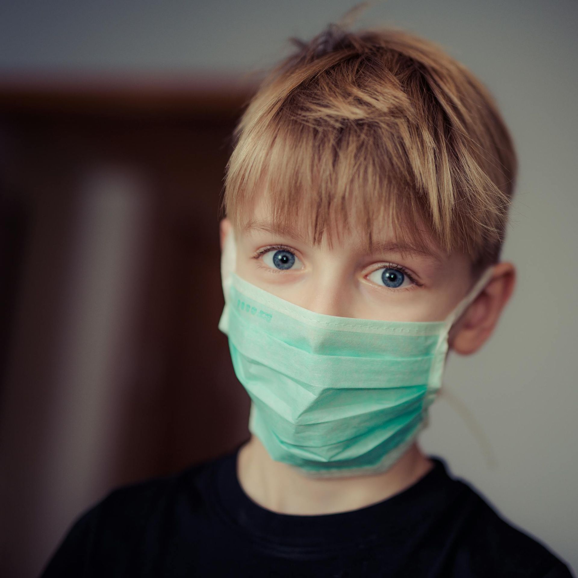 Blond boy with blue eyes wearing a green surgical mask indoors.