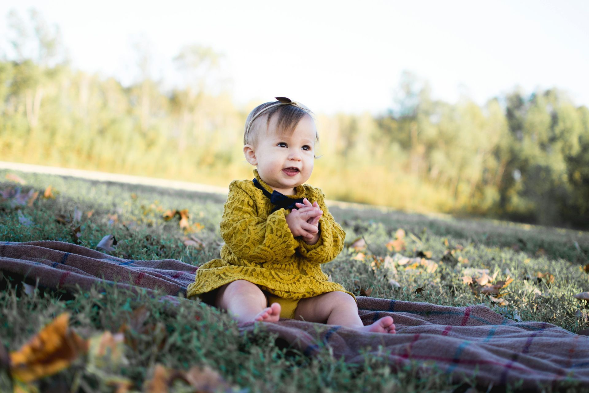 Baby girl in yellow dress sits smiling on a blanket in a grassy field.