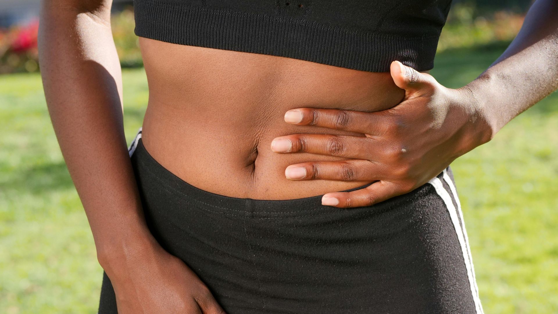 Black woman in athletic wear holding her hand on her abdomen, possibly indicating pain, in a sunny outdoor setting.