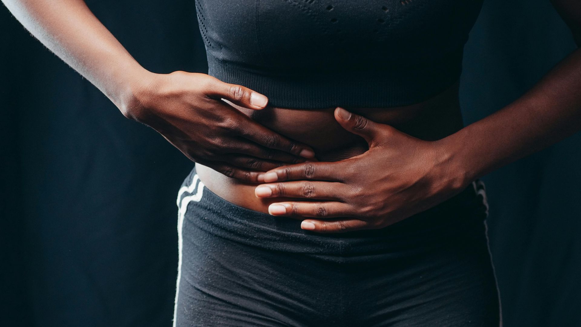 Woman in black sportswear, holding abdomen in pain against a dark background.