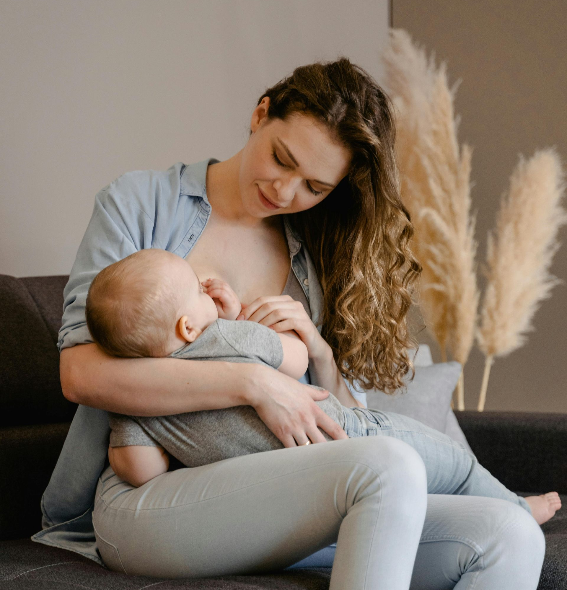 Woman breastfeeding baby on a couch, both looking down. Soft natural light, neutral tones.