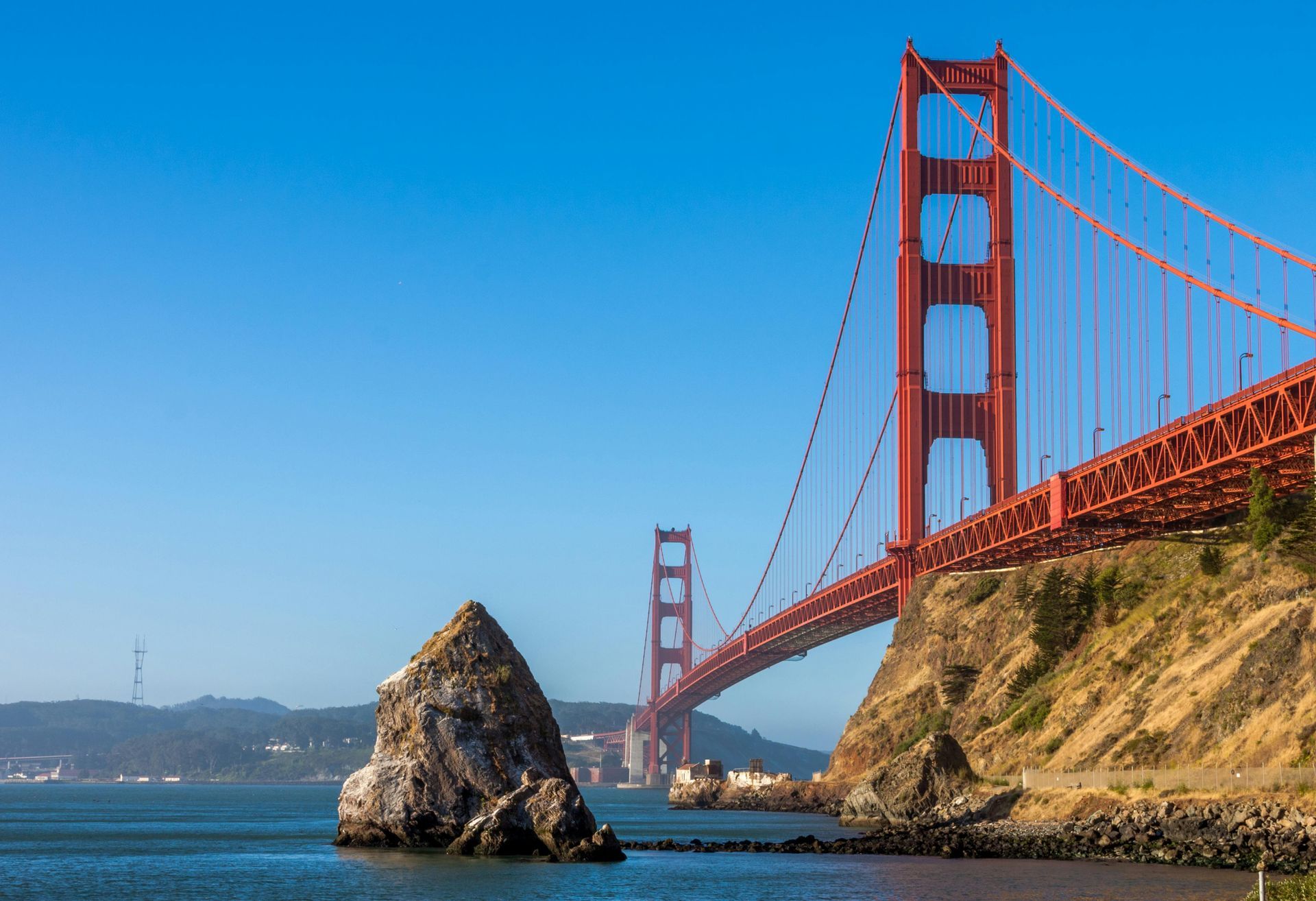 Golden Gate Bridge, red structure, spanning water, with rocky coastline and blue sky.