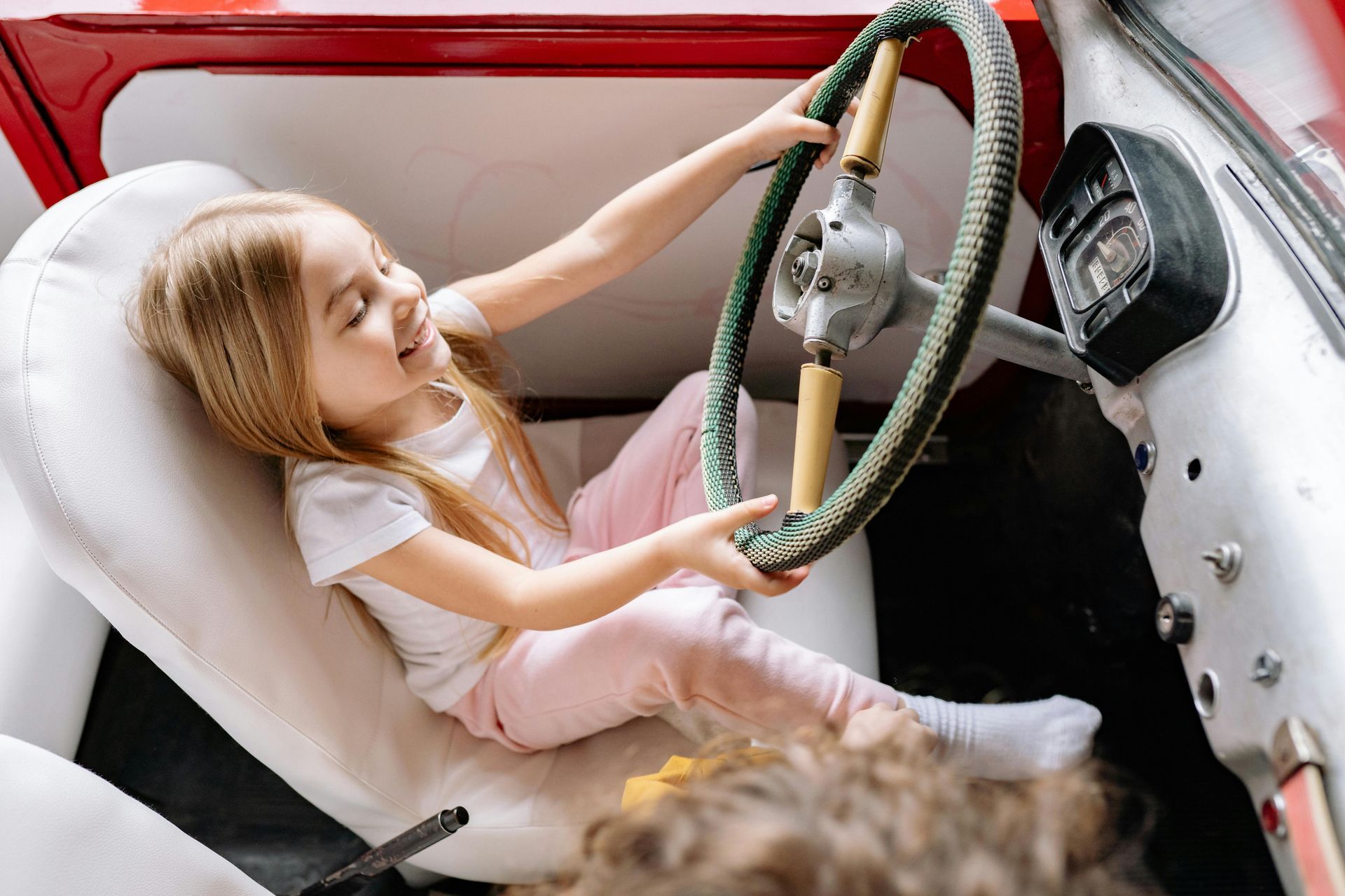Young girl in a white car seat, smiling and playing with the steering wheel.