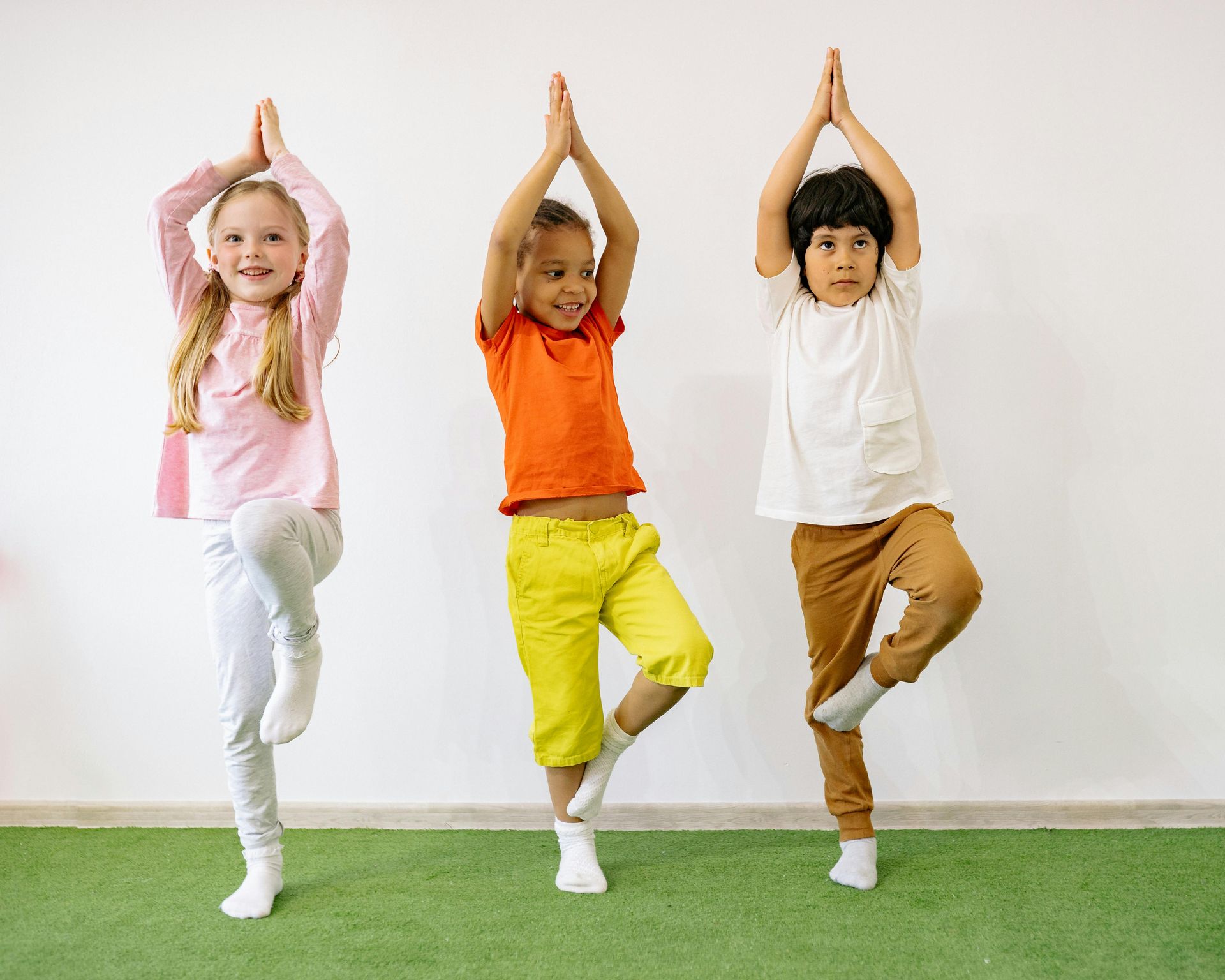 Three children of diverse ethnicities doing tree pose yoga in a brightly lit room.