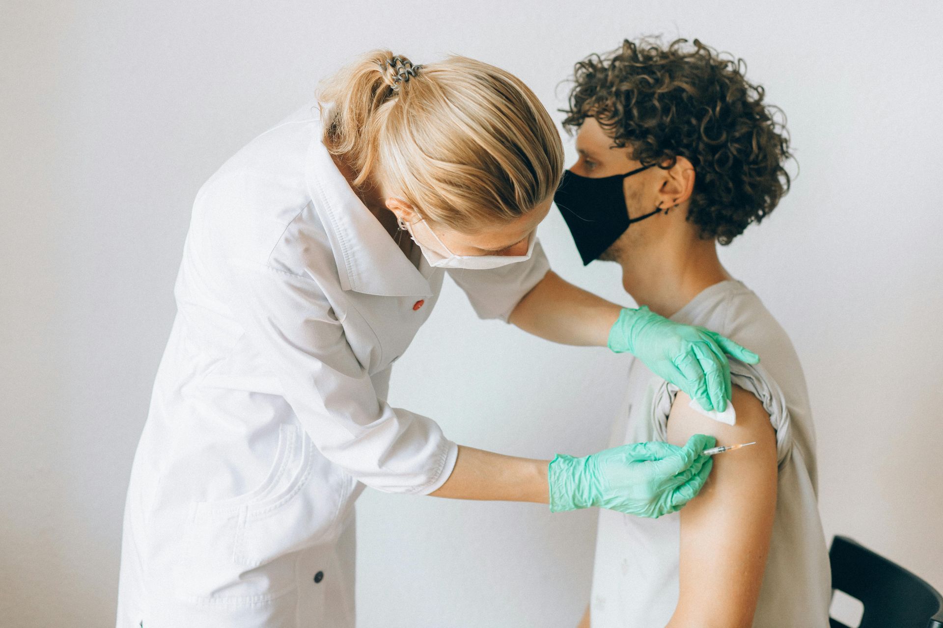 A healthcare worker giving a vaccine to a person wearing a mask. White coat, gloves, neutral background.