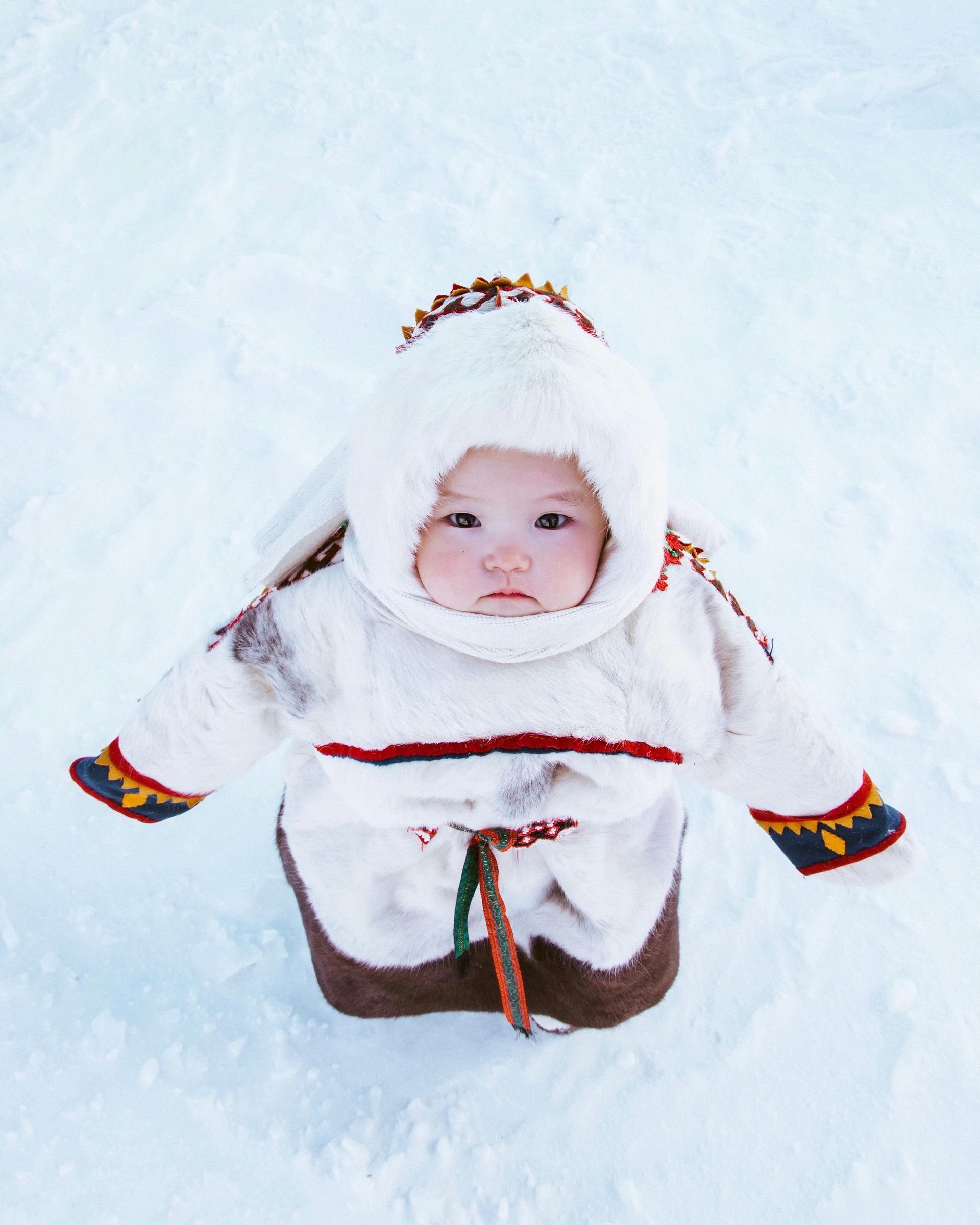 Baby bundled in white fur outfit sits in snow, looking up.