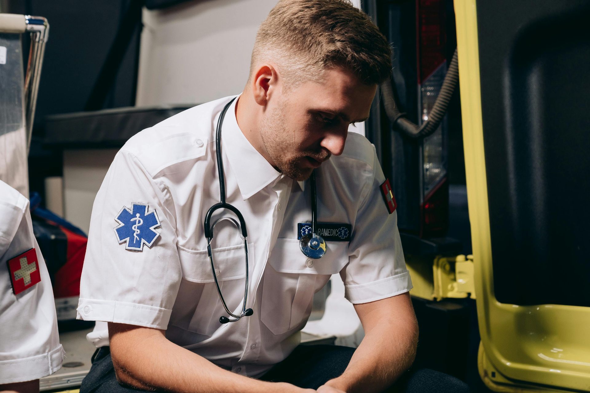 Paramedic in uniform with stethoscope, seated in an ambulance, looking down with a serious expression.
