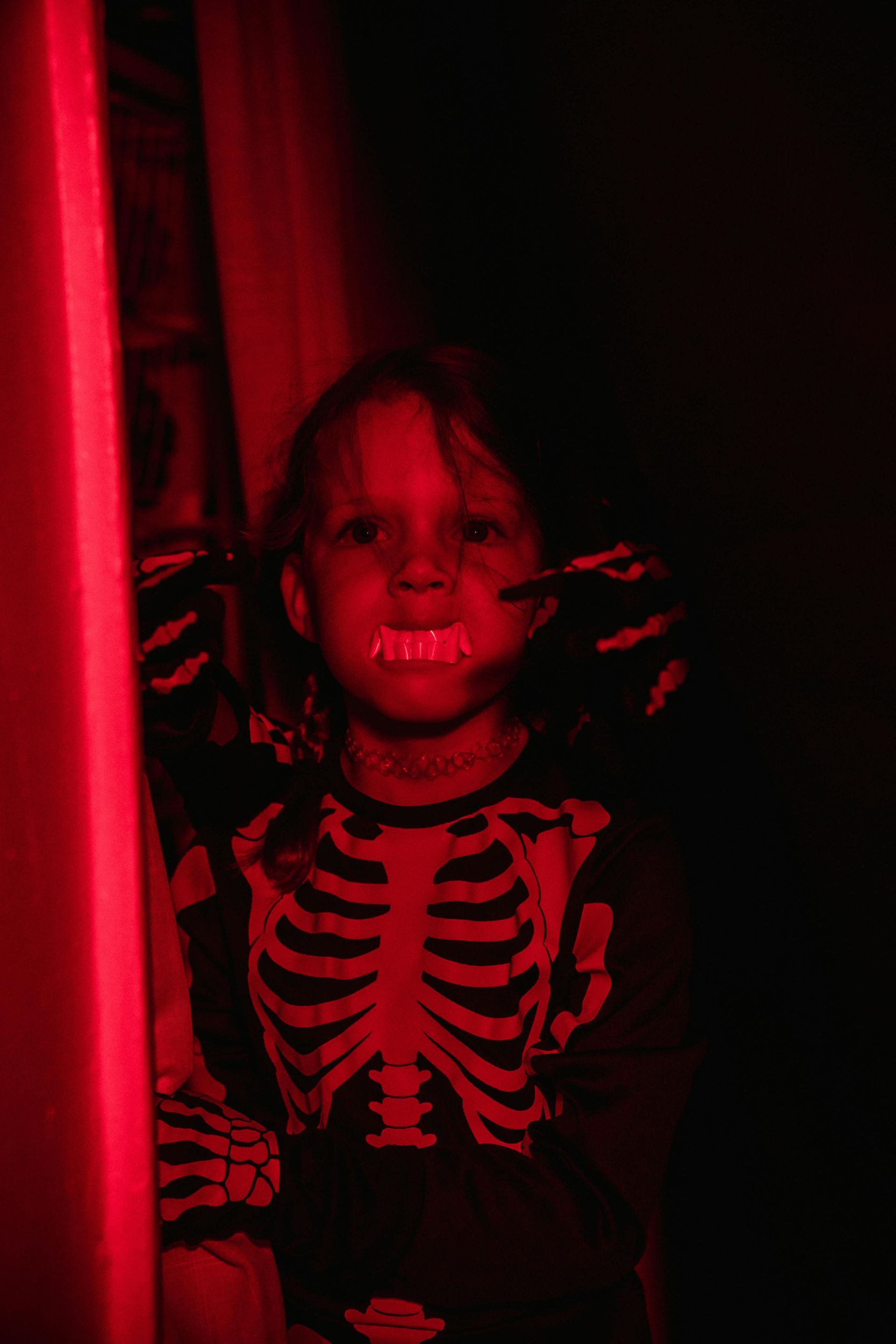 Child in skeleton costume with glowing teeth, red light illuminates face and surrounding dark space.
